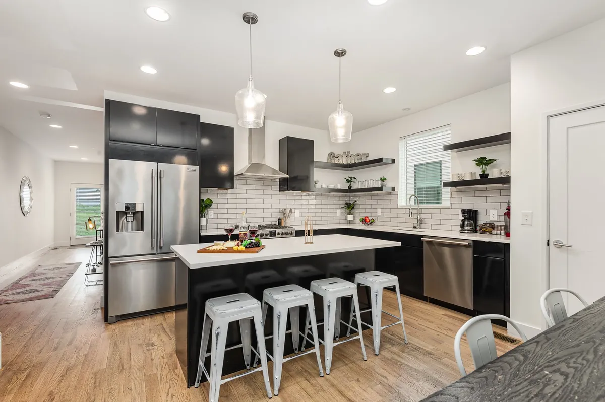 A modern, open-concept kitchen featuring a spacious island countertop, stainless steel appliances, sleek hanging light fixtures, and open shelving with decorative plants. The setting feels both functional and inviting, with plenty of room for multiple people to gather, cook, and chat.