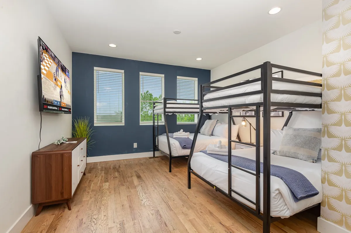 A playful bedroom featuring twin-over-twin and twin-over-full bunk beds, a large window letting in natural light, a television for group entertainment, and a wooden dresser. The space feels designed for group hangouts, movie nights, and sleepovers.
