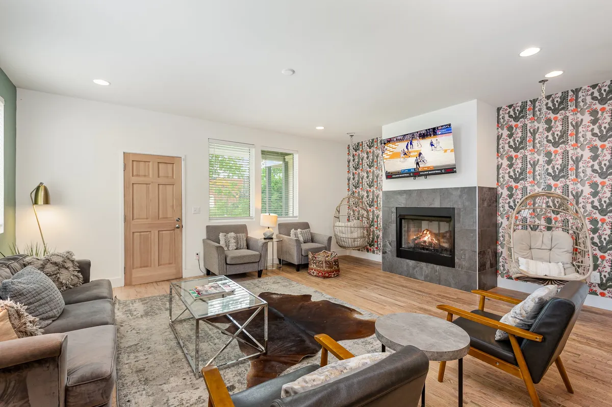 A cozy living room scene centered around a modern fireplace, with a comfortable seating area and a wall-mounted TV. The room is styled for relaxation and easy socializing, featuring warm tones and a hanging chair for a playful touch.