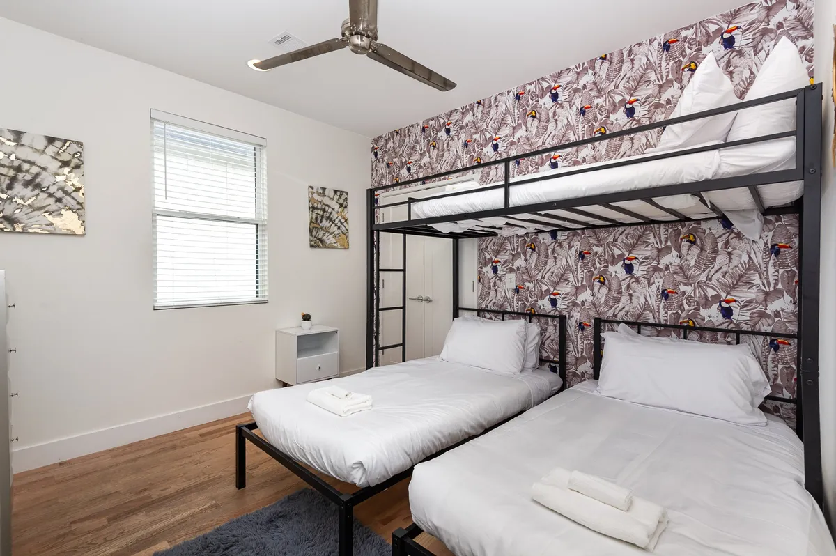 A cheerful bedroom showcasing a bunk bed and twin beds, accented by colorful wallpaper and a ceiling fan. The room is bright and inviting, ideal for kids or friends to share memories and enjoy sleepovers together.