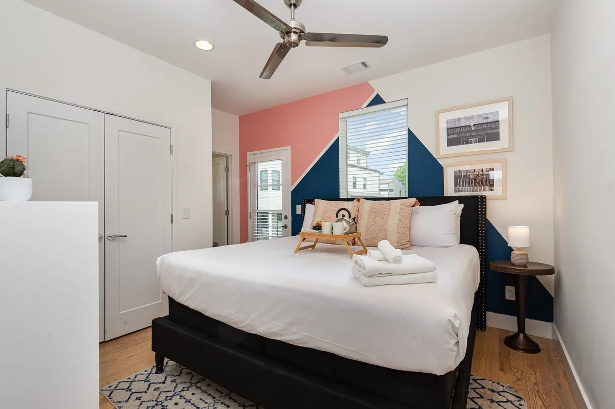 A cozy bedroom with a colorful accent wall, plush bed, decorative pillows, and framed artwork. Natural light streams in, creating a welcoming and relaxing environment.