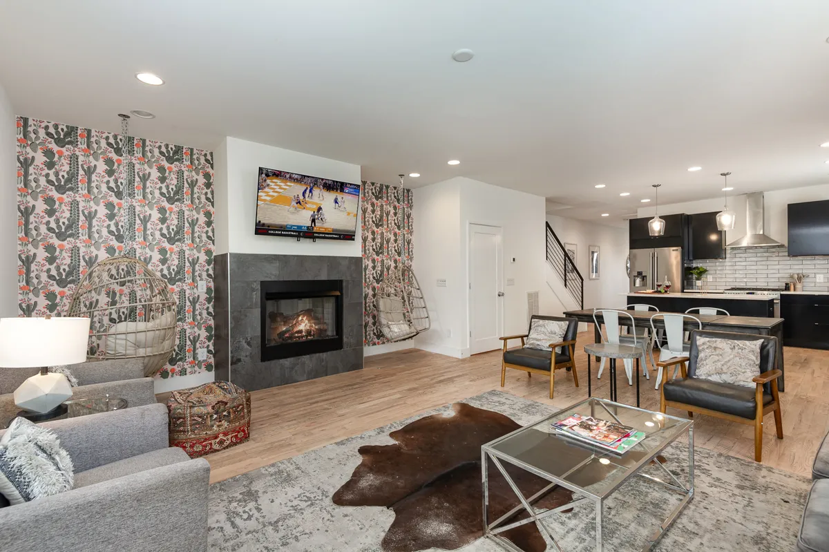 The image showcases a stylish, open-concept living room with a modern fireplace, vibrant wallpaper, comfortable seating including unique hanging chairs, a large smart TV above the mantel, and a sleek coffee table. The space feels warm, inviting, and perfect for groups to gather, relax, or watch a game together.